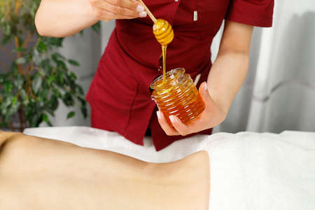 Close up photo of a jar of honey holding masseur in red uniform for a massage procedure to a young woman lying on the table in spa salon.の写真素材