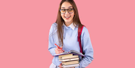 Excited school girl in uniform wearin glasses with backpack holding books and writing some notes in a diary on pink background.の写真素材