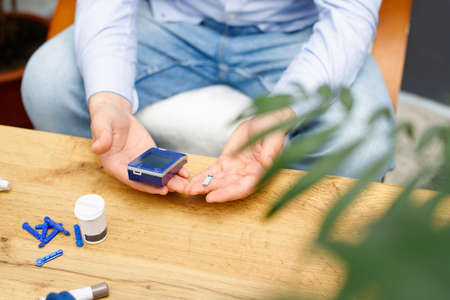 Man using glucometer and stripe for checking blood sugar level at home sitting on the chair at the table, diabetes concept.の写真素材