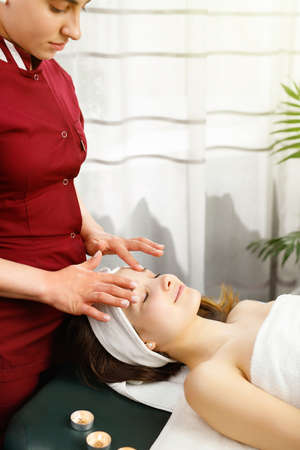 Beautiful woman enjoying facial massage with closed eyes in sunny spa center, masseur wearing red uniform.の写真素材