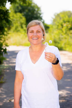 Picture of a happy sporty woman standing on the road in the forest wearing aid and digital watch, resting after jogging and holding the dandelion in one hand.の写真素材