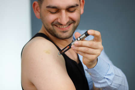 Smiling man making insulin injection to himself at home using syringe and being dressed part body with t-shirt, diabetes concept.の写真素材