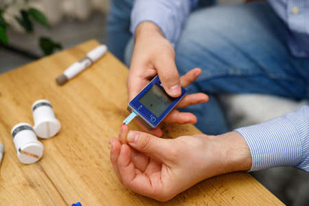 Close up photo of a single man measuring blood sugar level by glucose meter at home sitting on the sofa.の写真素材