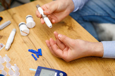 Top photo of a man sitting at the table at home using lancet on finger for checking blood sugar level by glucose meter, diabetes concept.の写真素材