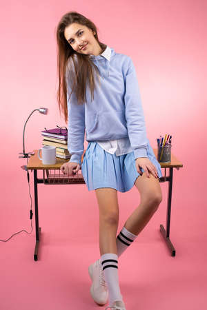 Student girl with blond long hair in school uniform and sneakers leaned on the table in the class posing for the camera on pink background.の写真素材