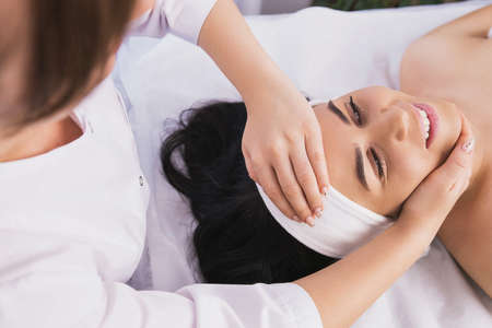 Above photo of cosmetologist making facial beauty massage treatment for relaxed young woman face with fingers movements in beauty spa salon.の写真素材