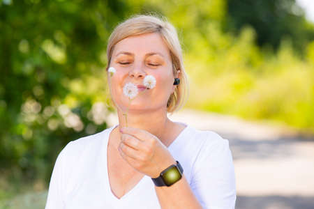 Close up photo of a happy blonde woman outdoors wearing digital watch and head phones holding in her hand the dandelion flower and is smiling.の写真素材