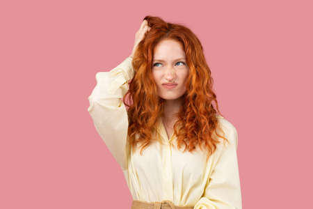 Picture of a thoughtful redhead young woman in yellow tunic put her hand on a head and looking up aside on pink background.の写真素材