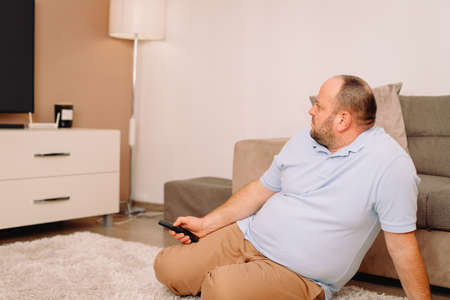 Side view photo of a sibgle man sitting on the carpet at home near the sofa holding in his hands tv remote and watching tv.の写真素材