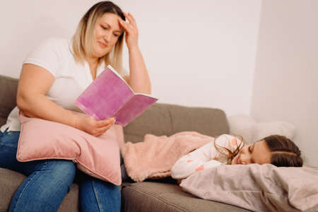 Mother reading to her child daughter book of pink color sitting on the sofa while little daughter sleeping.の写真素材