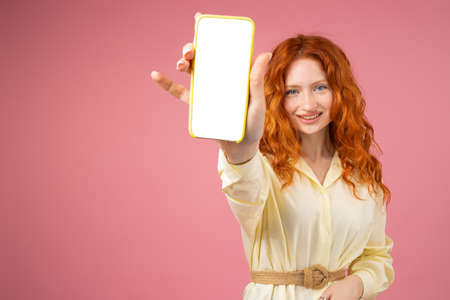 Picture of a happy redhead curly girl in yellow dress with a belt showing on camera phone with white screen in her hands over a pink background.の写真素材