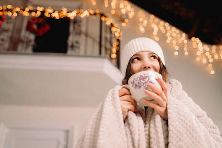 Beautiful young girl wearing white hat on her head and with blanket holding a cup of hot tea outdoors looking up in the sky.の写真素材