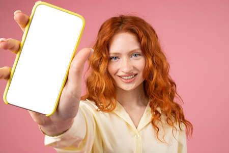 Redhead beautiful lady smiling in yellow clothes showing mobile phone on camera with white screen standing over pink background.の写真素材