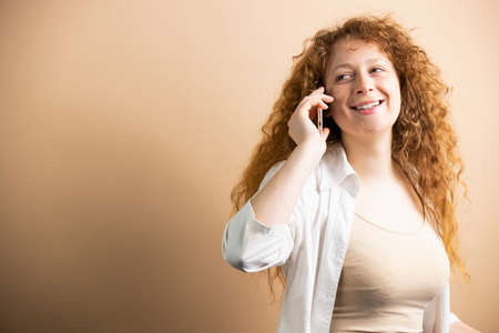 Modern pretty young woman with red long curly hair holding smartphone near the ear and talking with happy facial expression looking back.の写真素材