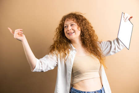 Funny young woman with red curly long hair holding hands aside and in one hand kepping tablet with white screen over golden background.の写真素材