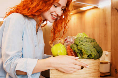 The beautiful red-haired girl looks at the basket of vegetables on the table.の写真素材