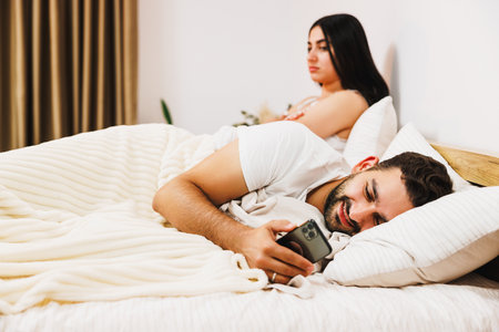 Young beautiful gorgeous sad girl sitting and hugging pillow on the bed in front of her boyfriend with mobile in hands.の写真素材