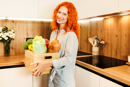 Young girl with red hair smiling at camera holding a wooden box of groceries from delivery.の写真素材