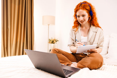 Young woman sitting on bed in bedroom and using laptop and make some notes.の写真素材
