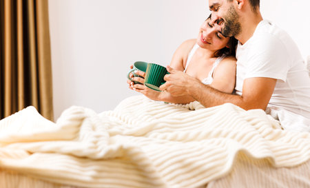 Happy caucasian couple smile and drink coffe together in bed in morning embracing each other.の写真素材