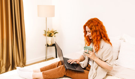 Smiling teen girl school student with orange hair use laptop computer sit on bed distance elearning online learning course prepare for exam search remote online course at home.の写真素材