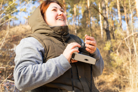 Joyful woman outdoors dressed in warm winter clothes keeping camera in hands and enjoying being in nature on sunny winter day.の写真素材