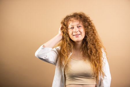Happy redhead young woman with long and wavy hair keeping her hand on head bending the arm at the elbow over golden background.の写真素材