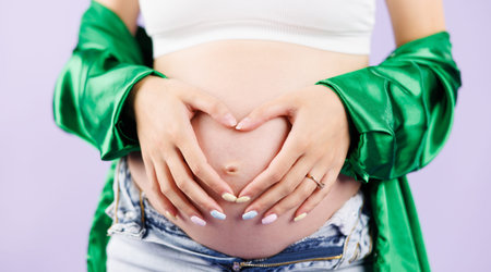Close up of pregnant woman in unzipped jeans holding her hands in a heart shape on her belly at...