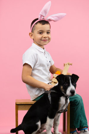 Full length photo of a little boy wearing bunny ears on head with basket ful of painted eggs sitting on a chair and a cute liitle dog near the boy.の写真素材