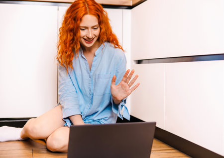 Happy smiling woman waving at her laptop computer as she interacts on a video conferencing call while sitting in an officeの写真素材