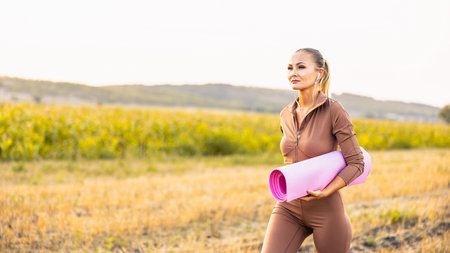 Young woman holds yoga mat in hand, running on the field. Healthy life style concept.の写真素材