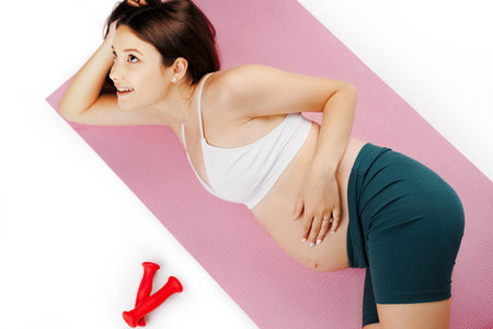 Relaxed beautiful pregnant with white top and green leggings women in yoga class lying on pink mats in a fitness studio with white background.の写真素材