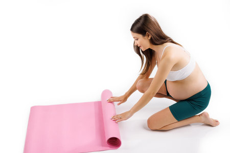 Beautiful and happy pregnant woman is preparing for gym exercises on the pink yoga mat on a white background.の写真素材