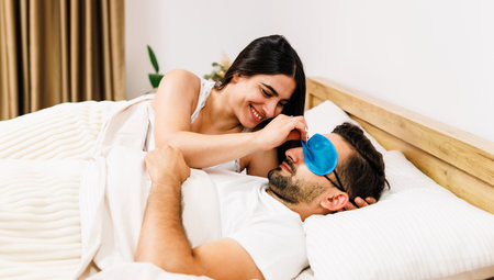 Young caucasian couple lying and covering under blanket on bed and she looking at he. Man a wear eye mask on face. Domestic lifestyle. Relationship. Modern bedroom.の写真素材
