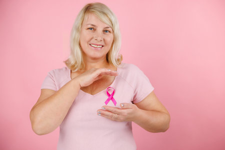 Woman holding pink cancer ribbon pointing thumb up to the side smiling happy with open mouthの写真素材