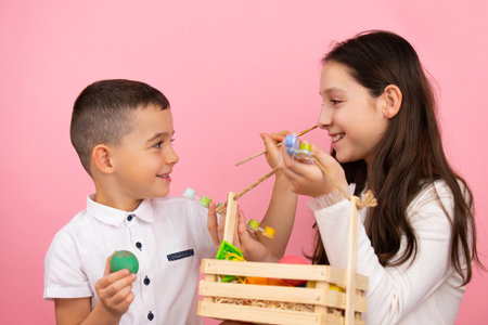 Side view photo of brother and sister preparing for the Easter holiday, he holds green egg and with brush painting the sister nose while she holds a basket with eggs.の写真素材