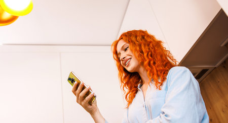 Young fun smiling housewife woman 20s wearing casual clothes striped t-shirt use mobile cell phone chatting online browsing surfing internet in light kitchen at home alone People lifestyle concept.の写真素材