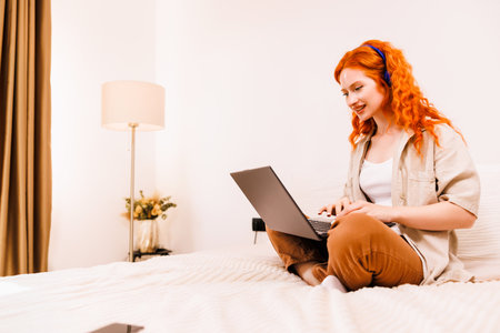 Pretty young woman sitting cross-legged on bed with laptop and class notes studying at home looking at computer with a smileの写真素材