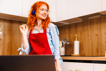 Smiling beautiful woman dancing in modern kitchen, preparing healthy food alone, cooking salad, carefree happy girl singing and moving to favorite music, having fun at home, enjoying leisure timeの写真素材