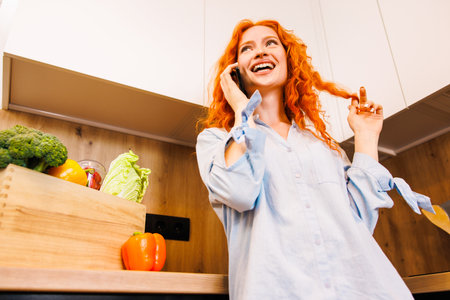 Happy joyful chef girl making telephone call in kitchen, cooking salad from fresh vegetables for dinner, talking on mobile phone at table with healthy food, smiling, laughing. Domestic work conceptの写真素材