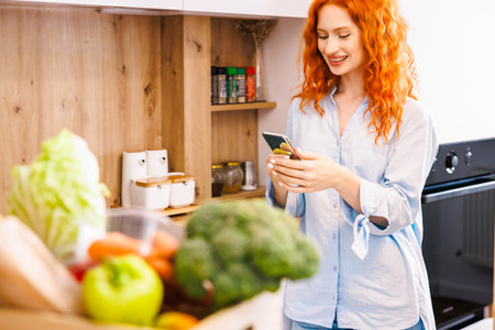 Young happy woman is holding white cup and looking at the camera while sitting at wooden table in the kitchen among green vegetablesの写真素材