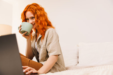 Beautiful young red haired smiling woman working on laptop and drinking coffee while sitting in a big comfortable couch covered with blanket over shoulders at homeの写真素材