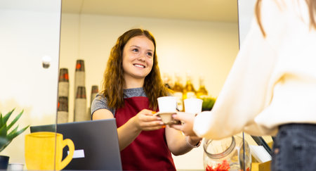 Best coffee to go. Part of young cheerful Caucasian woman in apron holding coffee cups while standing at cafe.の写真素材