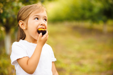 Beautiful girl eats fresh, ripe and tasty plum, close up. Kid bites delicious fruit and looks aside. Summer nutrition and healthy food conceptの写真素材