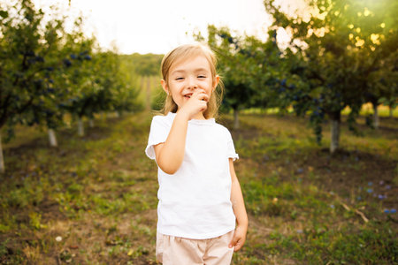 Cute little girl is eating plums in the orchard dressed in white clothes.の写真素材