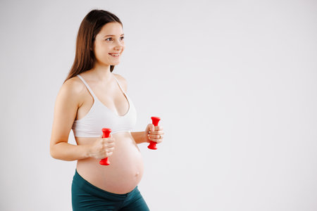 Happy Pregant woman with brown hair doing fitness exercise in white top and green leggings with red dumbbell isolated on white background.の写真素材
