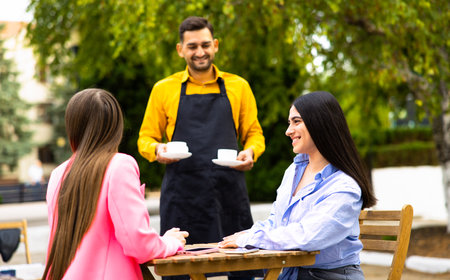 Smiling friendly waitress serving coffee drinks to diverse male friends meeting at cafe table, cafeteria server and multiracial visitors laughing together at funny joke in modern coffee shop houseの写真素材