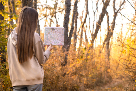 Back view of stylish woman holding paper map, wearing warm beige sweater and jeans, looking at...
