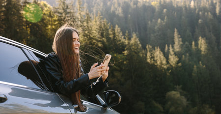 Young beautiful woman traveling by car in the mountains using smartphone at sunsetの写真素材