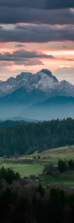Majestic snow-capped mountains are silhouetted against a dramatic sunset sky above rolling green...の写真素材